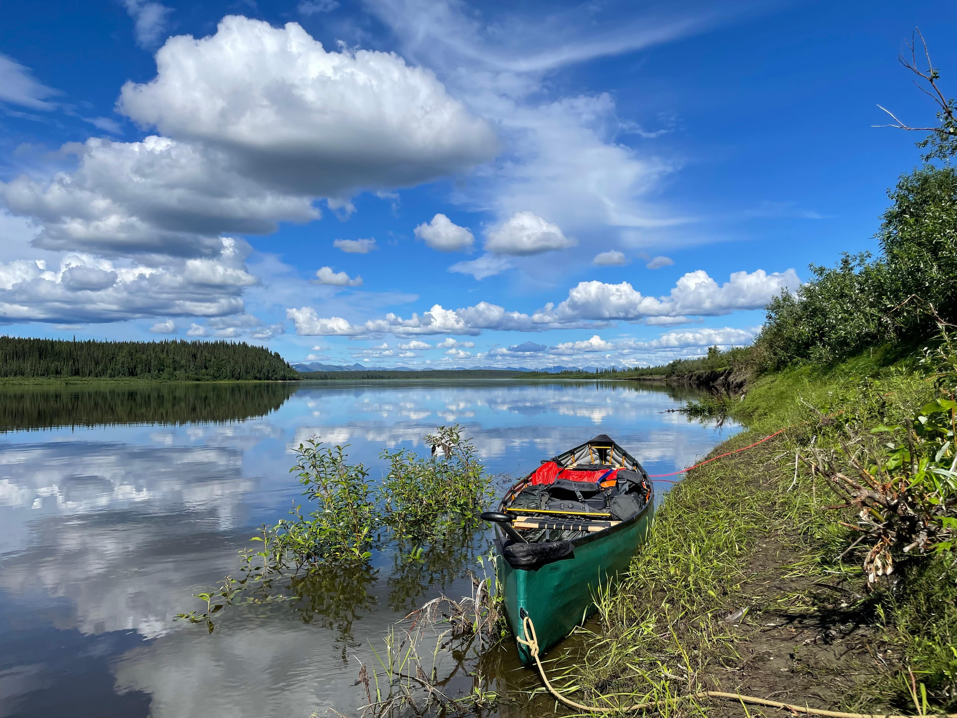 A green pack canoe sits on the bank of the Kobuk River. Clouds are mirrored on the water's surface.